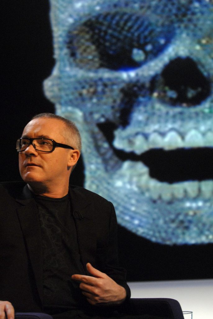 Damien Hirst in front of a photograph of For the Love of God, which he apparently still owns. (Photo by Jeff Overs/BBC News & Current Affairs via Getty Images)