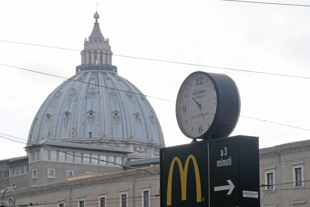 A sign showing the direction of a McDonald's restaurant is seen with the cupola of St. Peter's basilica in the background, on January 3, 2017 in Rome. The US fast-food chain opened a restaurant in a Vatican-owned building despite protest of a Committee for the Protection of Borgo, the historic district around the Vatican where many cardinals live. Photo by Tiziana Fabi for AFP via Getty Images.