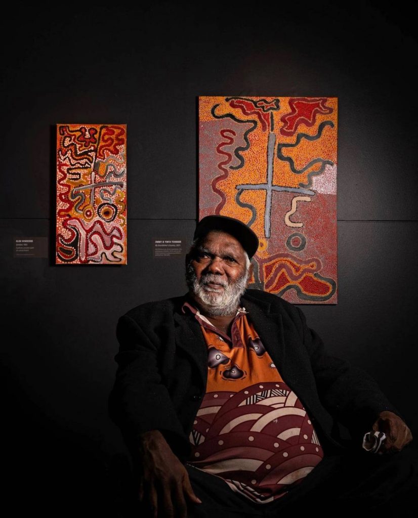 Balgo artist Jimmy Tchooga with the painting he created (right) with his wife Yintji in response to seeing his grandfather Alan Winderoo’s first painting from 1982 (left) at "Balgo Beginnings" at the South Australian Museum. Photo by Brad Griffin, courtesy of the South Australian Museum, Adelaide. 