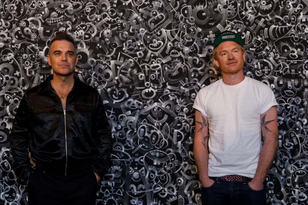 Robbie Williams (left) and Ed Godrich posing in front of their painting <i>Sharon</i> at their debut exhibition 'Black and White Paintings' at Sotheby's London. Courtesy Sotheby's. 