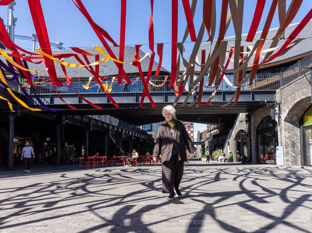 Sheila Hicks walking beneath 'Woven Wonders' her installation in Coal Drops Yard, King's Cross. Courtesy the artist and Alison Jacques Gallery.