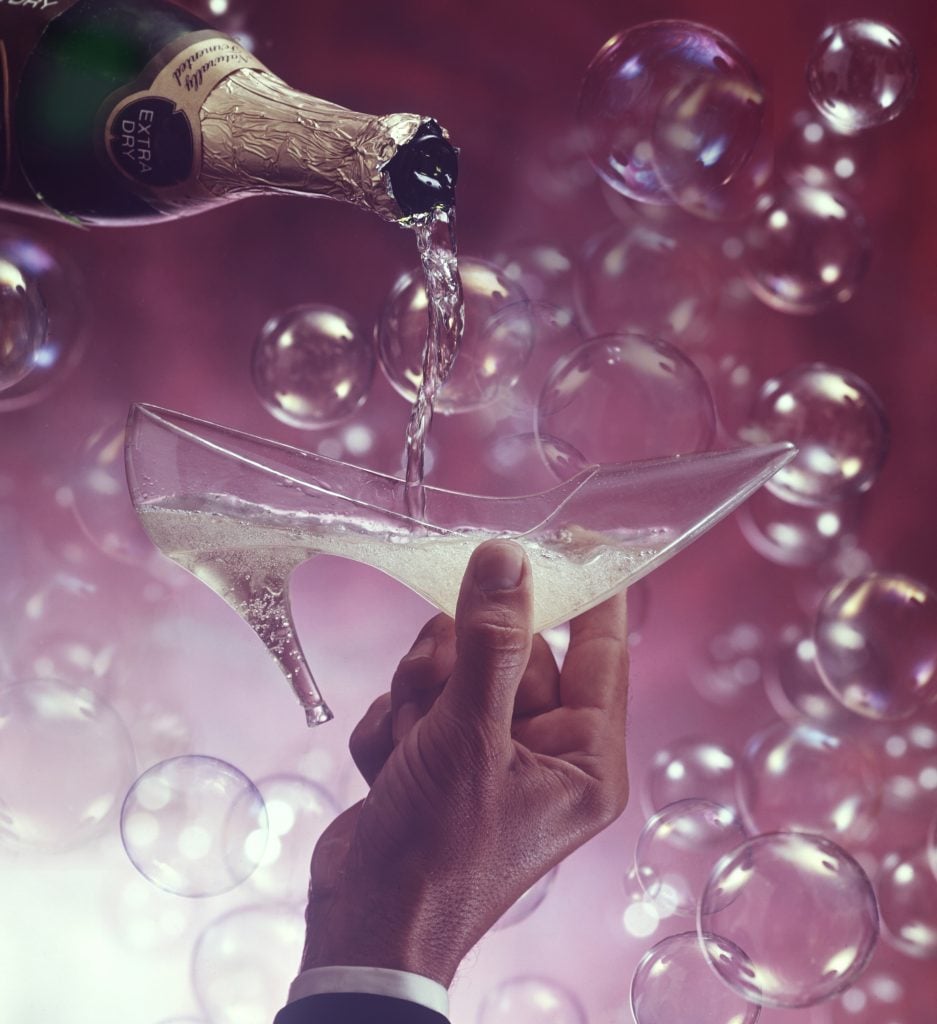 In a celebratory setting, a man's hands pour champagne into a glass slipper, while bubbles float in the background, ca.1970s.