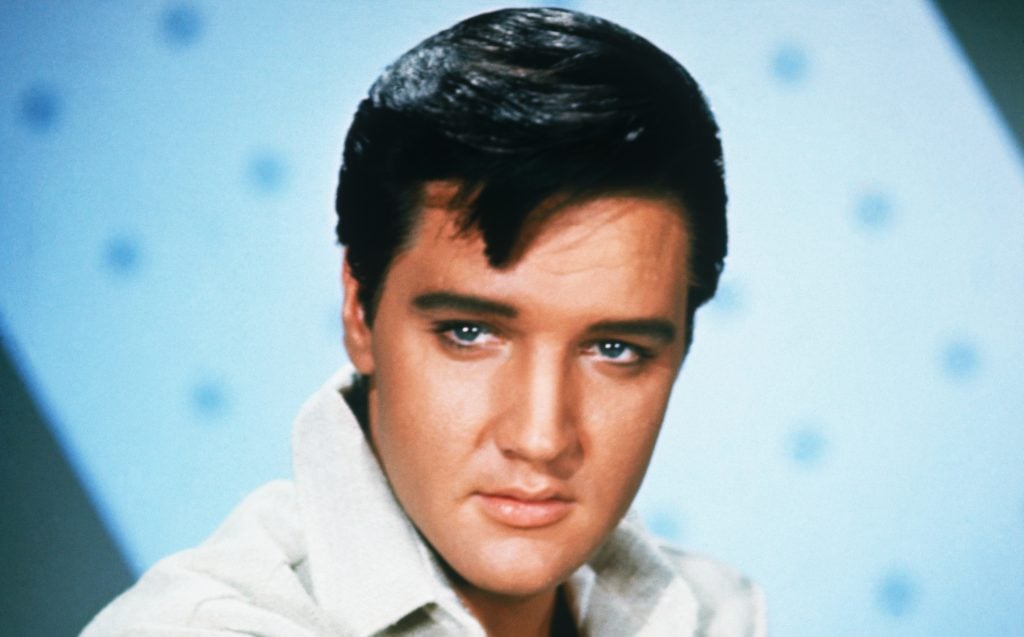 A headshot of a young Elvis Presley in a white colored shit, a lock of dark hair falling onto his forehead.