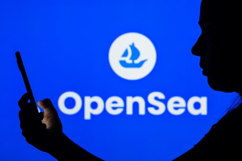 A woman's silhouette holds a smartphone with the OpenSea logo in the background. Photo: Illustration by Rafael Henrique/SOPA Images/LightRocket via Getty Images.
