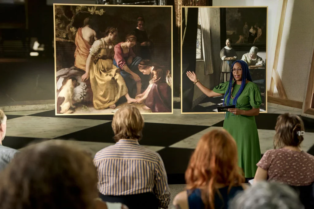Photo of a woman standing in front of an audience pointing to a Vermeer painting