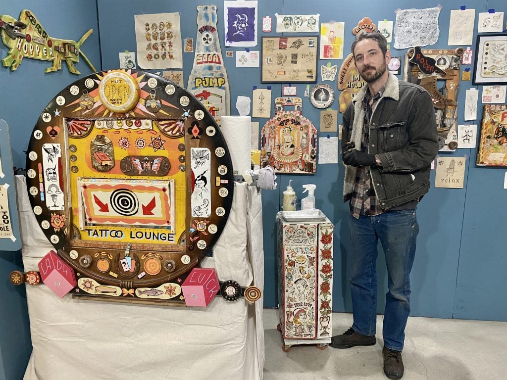 Kevin Hennessy with his work—and tattoo glory hole—at Spring Break Art Show Los Angeles 2023. Photo by Sarah Cascone.
