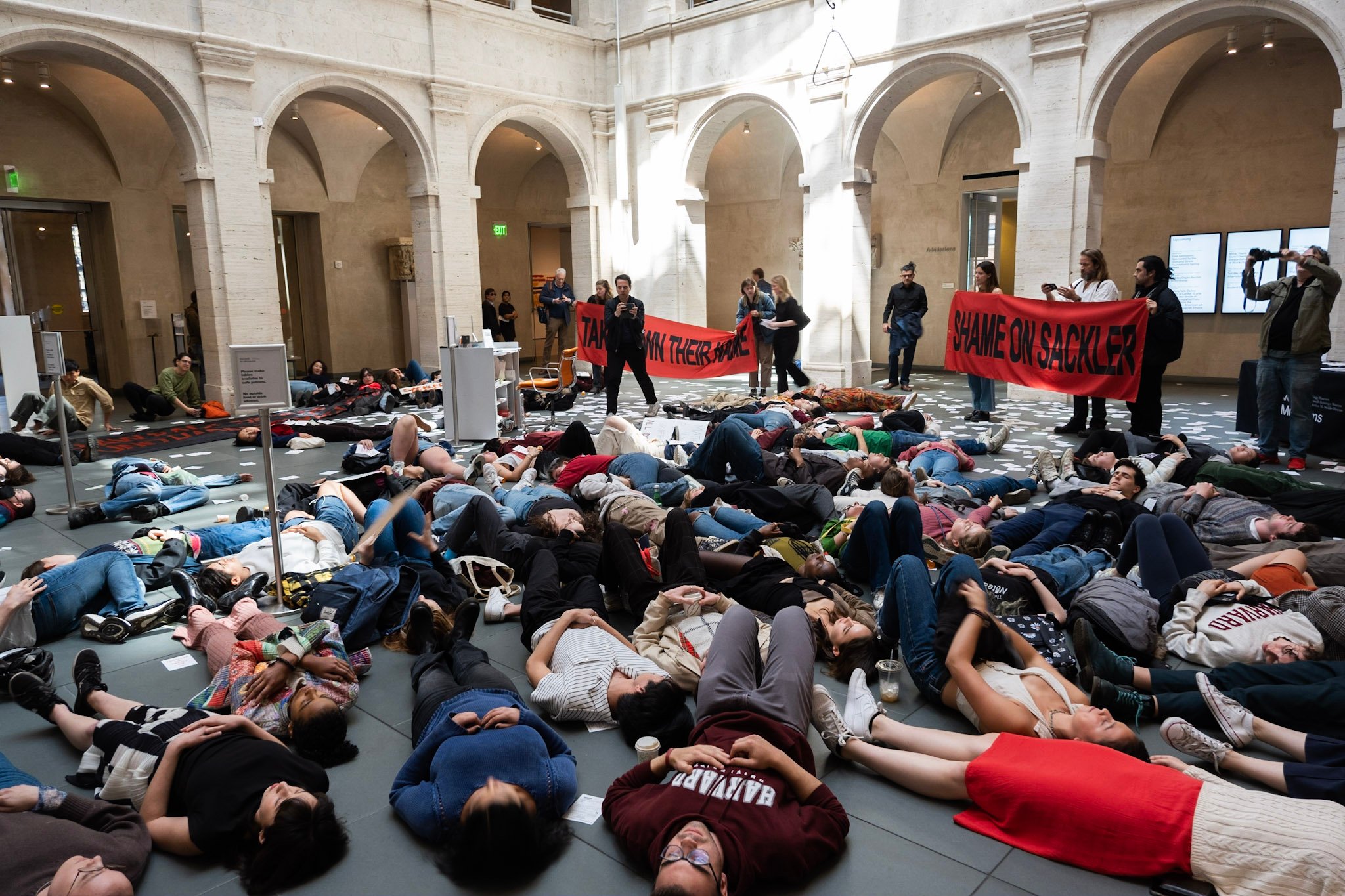 Protestors lie down in a 'die-in" in courtyard with an ornate arcade at Harvard's Arthur M. Sackler Museum. Some protesters stand holding red banners with the words "Shame on Sackler" and "Take Down Their Name" in black capitol letters.