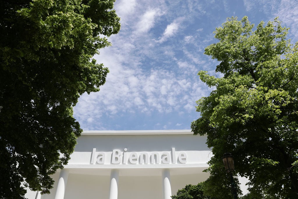 the image is of the top of a white building surrounded by trees and a blue sky with clouds in the background
