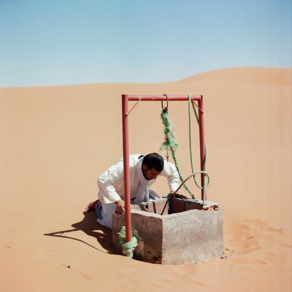 A man looking for water in a well surrounded by golden dunes at the oasis of Merzouga, Morocco, 2021, from the series ‘Before it’s gone
