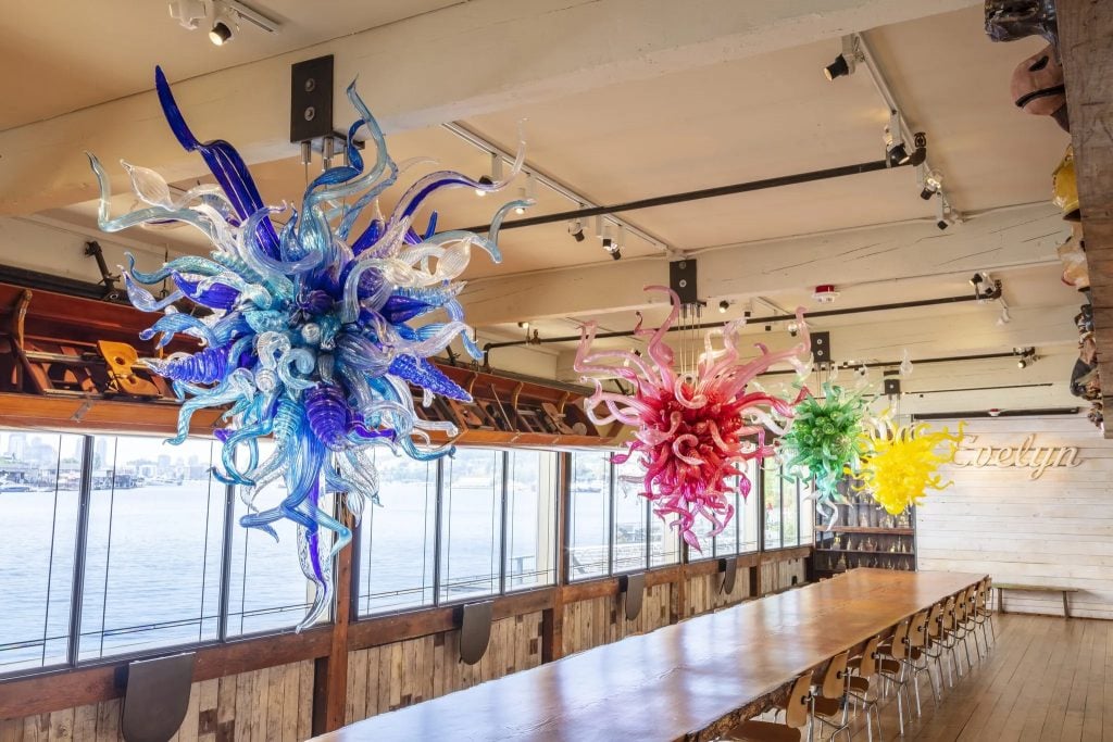 Dale Chihuly chandeliers and an Algonquin Tribe hand-carved birch canoe over a 85-foot-long Douglas fir table in the Evelyn Room at the Boathouse, the artist's Seattle studio. Photo by Nathaniel Willson, ©2023 Chihuly Studio.