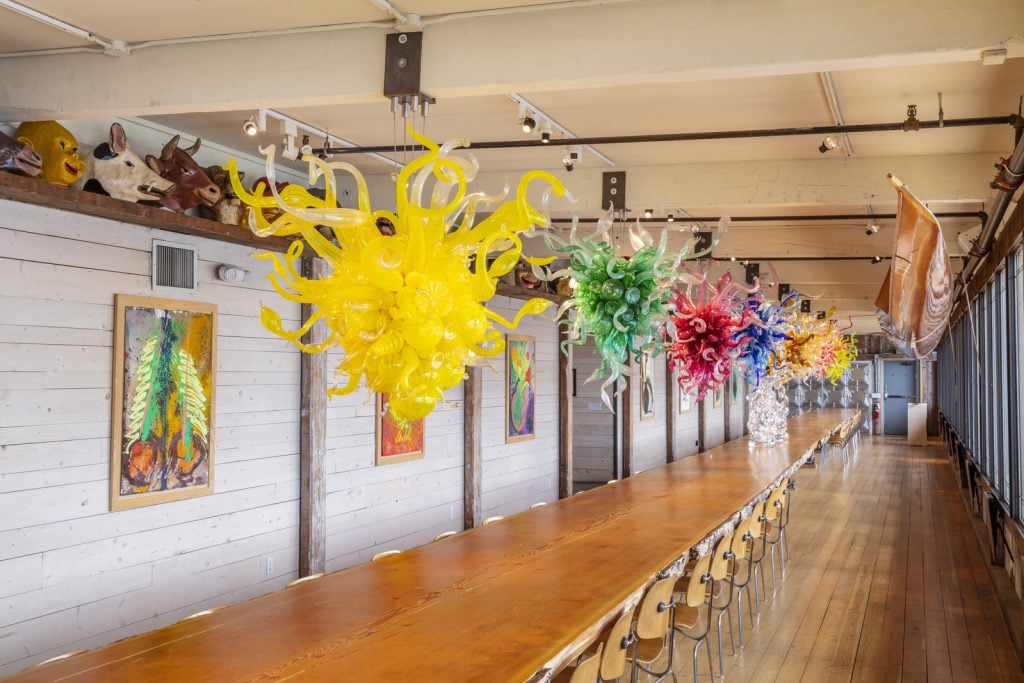 Dale Chihuly chandeliers and paintings, as well as giant papier-mâché masks and an Algonquin Tribe hand-carved birch canoe, over a 85-foot-long Douglas fir table in the Evelyn Room at the Boathouse, the artist's Seattle studio. Photo by Nathaniel Willson, ©2023 Chihuly Studio.