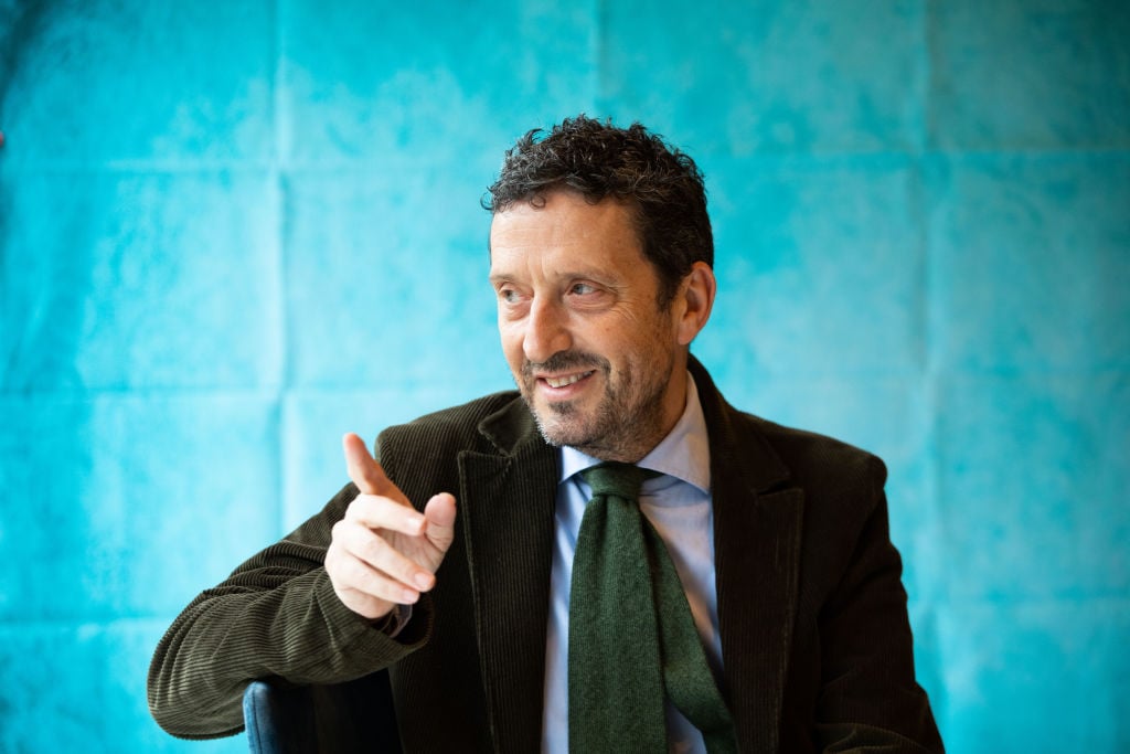 a smug looking man wags his finger against a light blue background