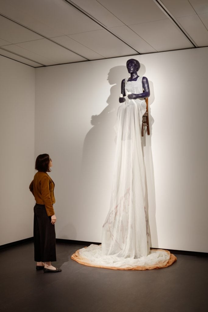 Alison Saar's <em>Undone</em> (2012) in "The Sky's the Limit" at the National Museum of Women in the Arts. Photo by Jennifer Hughes, courtesy of NMWA. A museum visitor gazes at a tall sculptural work hanging from the wall. The sculpture is of a dark skinned woman seated in a dark chair. The figure wears an elongated sheer white dress with a pink hem that extends far below the bottom of the chair.