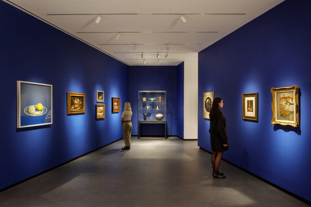 Collection galleries at the National Museum of Women in the Arts. Photo by Jennifer Hughes, courtesy of NMWA. Museum visitors observe artworks hanging on the royal blue walls of a small gallery bay. Paintings of various sizes are on display, and small sculptures are in a glass case.