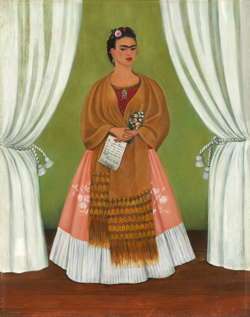 Frida Kahlo, Self-Portrait Dedicated to Leon Trotsky (1937). Collection of the National Museum of Women in the Arts, gift of Clare Boothe Luce; ©Banco de México Diego Rivera Frida Kahlo Museums Trust, Mexico, D.F./Artists Rights Society (ARS), New York; Image by Google. In a painted self-portrait, the artist stands in a stage-like space framed by white curtains. Beneath black hair woven with red yarn and flowers, heavy brows accent her dark-eyed gaze. Clad in a fringed, honey-toned shawl; long, pink skirt; and gold jewelry, she holds a bouquet and a handwritten letter.