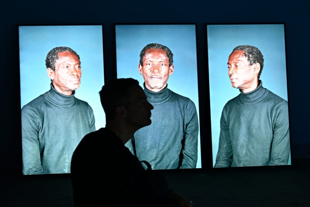 An art exhibit visitor is silhouetted against three large, portrait-oriented screens displaying the same image of a man in a dark turtleneck. The man’s poses vary slightly: he faces forward with a neutral expression, looks directly at the camera with a subtle smile, and turns his profile to one side. The images have a bright, high-contrast quality, and a cool-toned background highlights the subject.