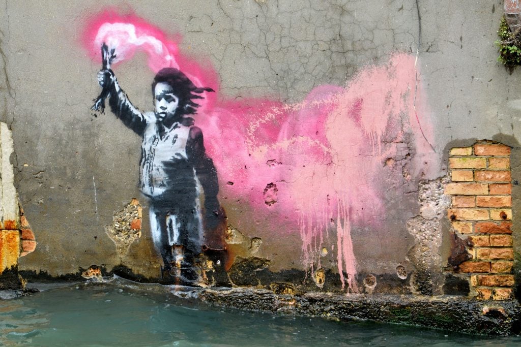 A Banksy mural, depicting a migrant child wearing a lifejacket holding a pink flare, is painted on the outer wall of a house overlooking the canal Rio de Ca Foscari, in Venice. Photo by Marco Sabadin / AFP.