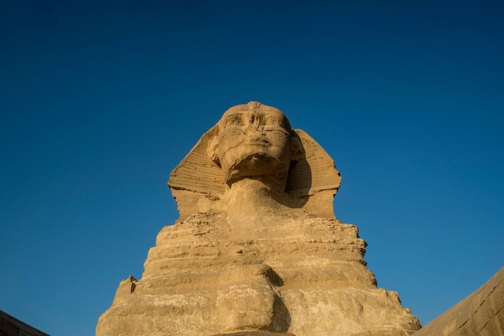 The face of the Great Sphinx of Giza, without a nose. Pictured against a blue sky.