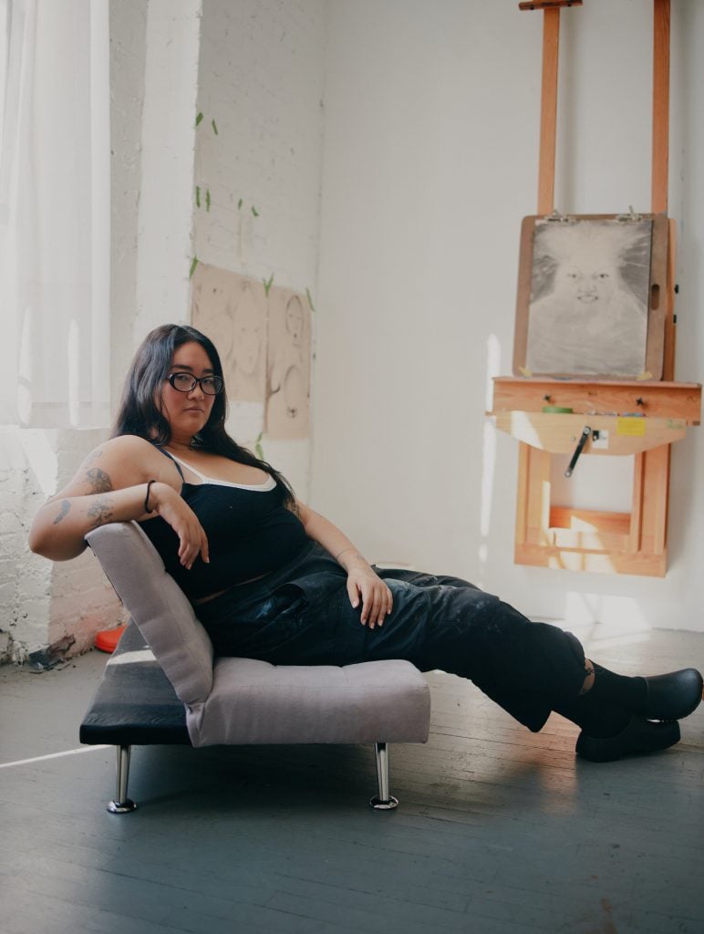 Contemporary artist reclining in her studio with sketch portraits and an easel in the background, highlighting creative process and modern art practice.