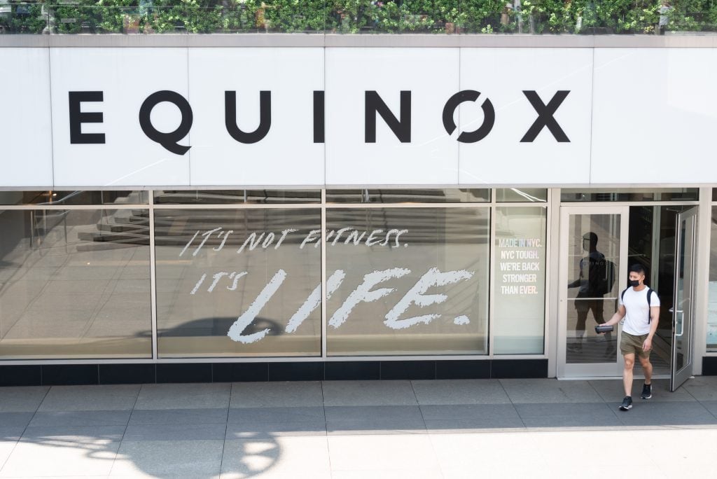 A few people walk out of a store with a big sign reading EQUINOX on a sunny day. They are in workout clothes.