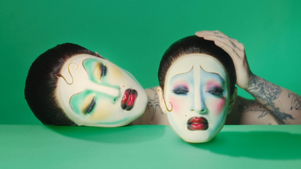 a person in makeup holding a doubble of her face in the same makeup against a green background
