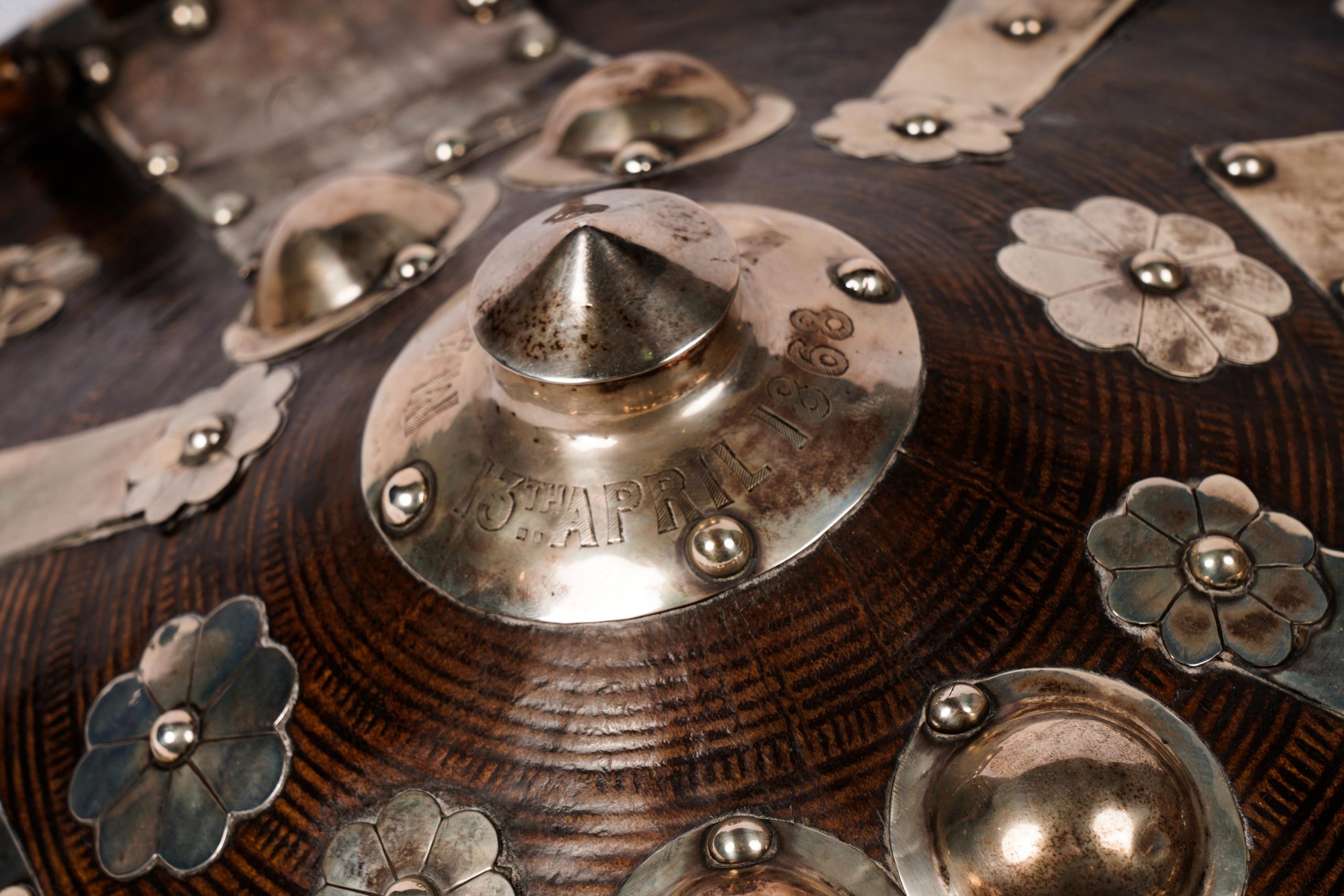 The shield in the image is a traditional Ethiopian (Abyssinian) shield from the year 1868. It features a convex, circular shape and is constructed from a dark, hardened leather. The surface is adorned with metal accents, which are both decorative and functional, serving to reinforce the shield's structure. The metal work displays a series of patterns and motifs, including flowers and embossed circular designs that add to its aesthetic appeal.