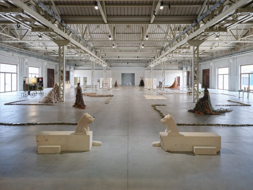 Art installation depicting sphynxes and other sculptures made with natural materials like ceramics and clay, in a large industrial space