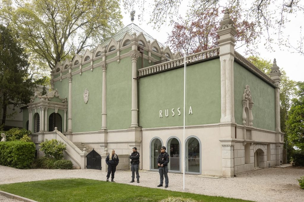 Security guards stand outside of a Neoclassical building painted light green building with lettering that identifies it as the Russia pavilion at the Venice Biennale. The building is surrounded by trees and a small grass lawn.