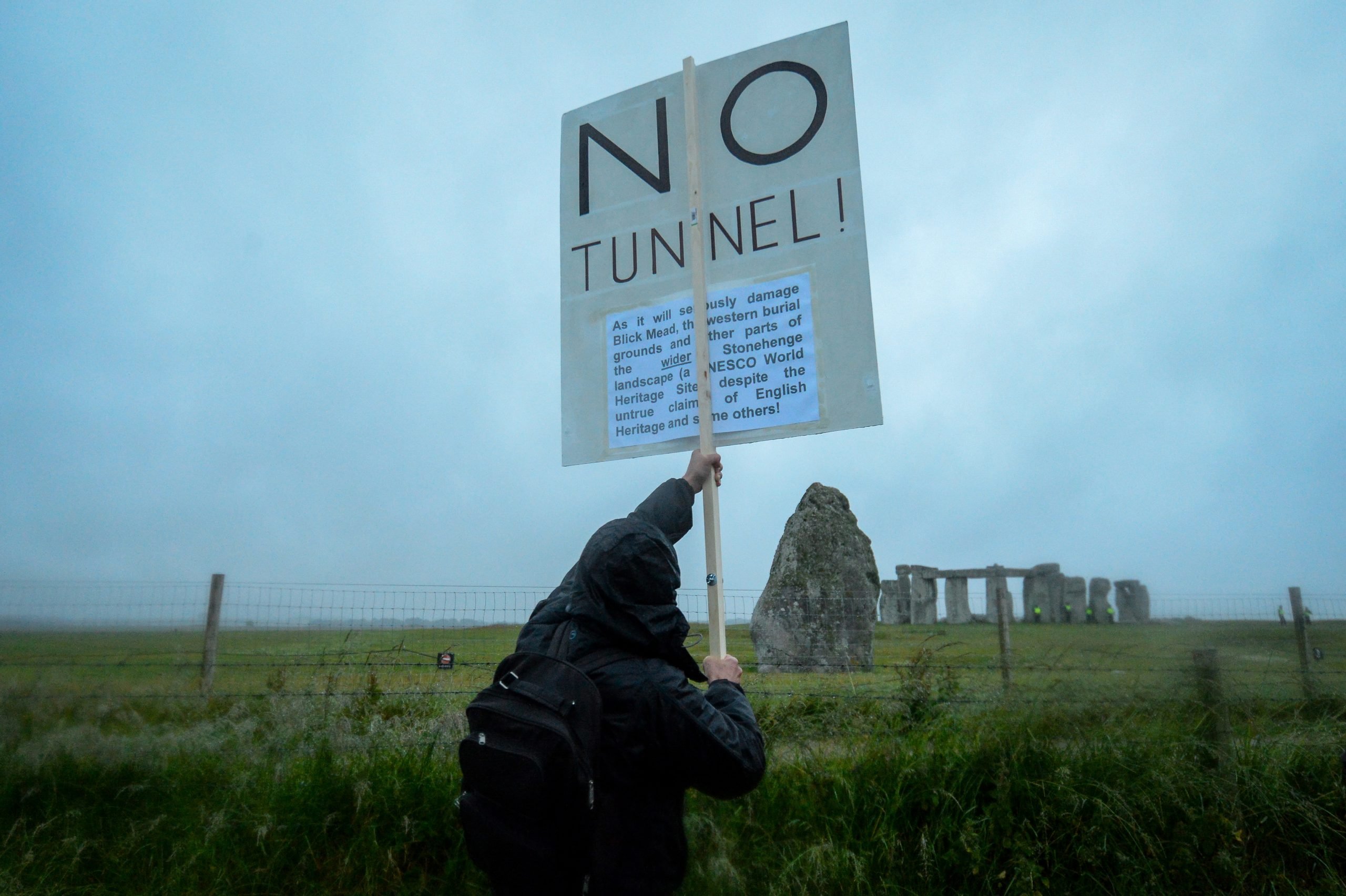 a person holding a protest sign stands in front of the stonehenge monument in england. the sky is grey and rainy