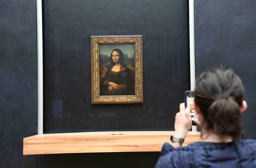 In a color photo, a person is seen from behind, using their camera phone to take a photograph of a painting of a woman in a gold frame.