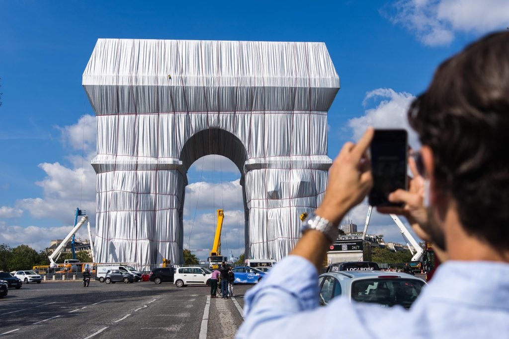 a man takes a cell phone picture of the arc de triomphe wrapped in fabric