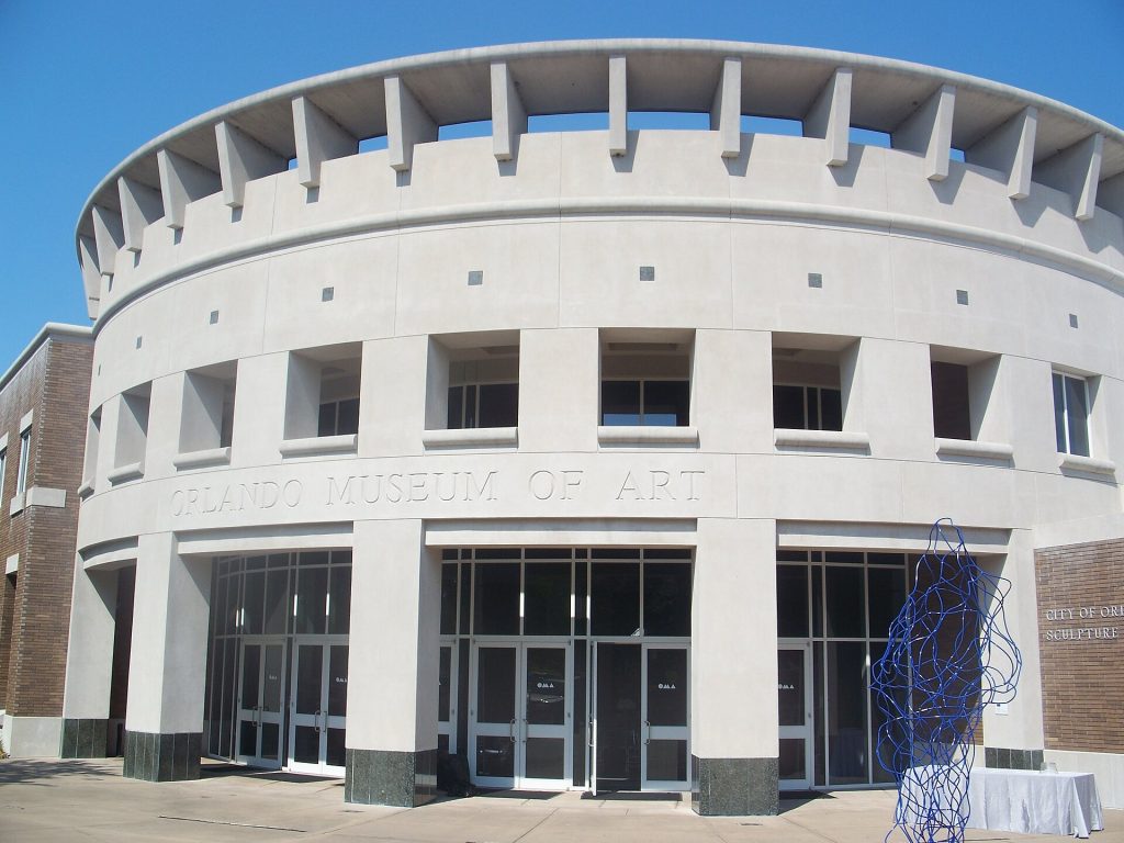 A color photo shows a cylindrical building of a few stories. The sky is blue behind it.