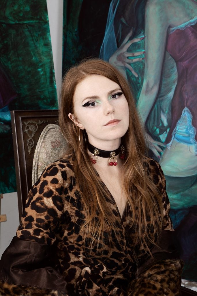 A white woman with long auburn hair looks out at the camera. She wears a black choker on her neck and blue eyeshadow with winged eyeliner.