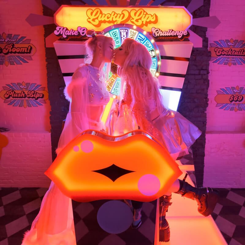 The image shows two individuals in a vibrant and colorful setting, reminiscent of an amusement arcade or themed party room. They are dressed in whimsical clothing with elements that sparkle or reflect light, giving a futuristic or fantasy-like appearance. Both have long blonde hair and are facing each other, about to kiss. In the foreground, there's a large, bright orange, lip-shaped object, a part of an interactive game at the Museum of Sex’s Superfunland. The background is lit with neon signs saying 