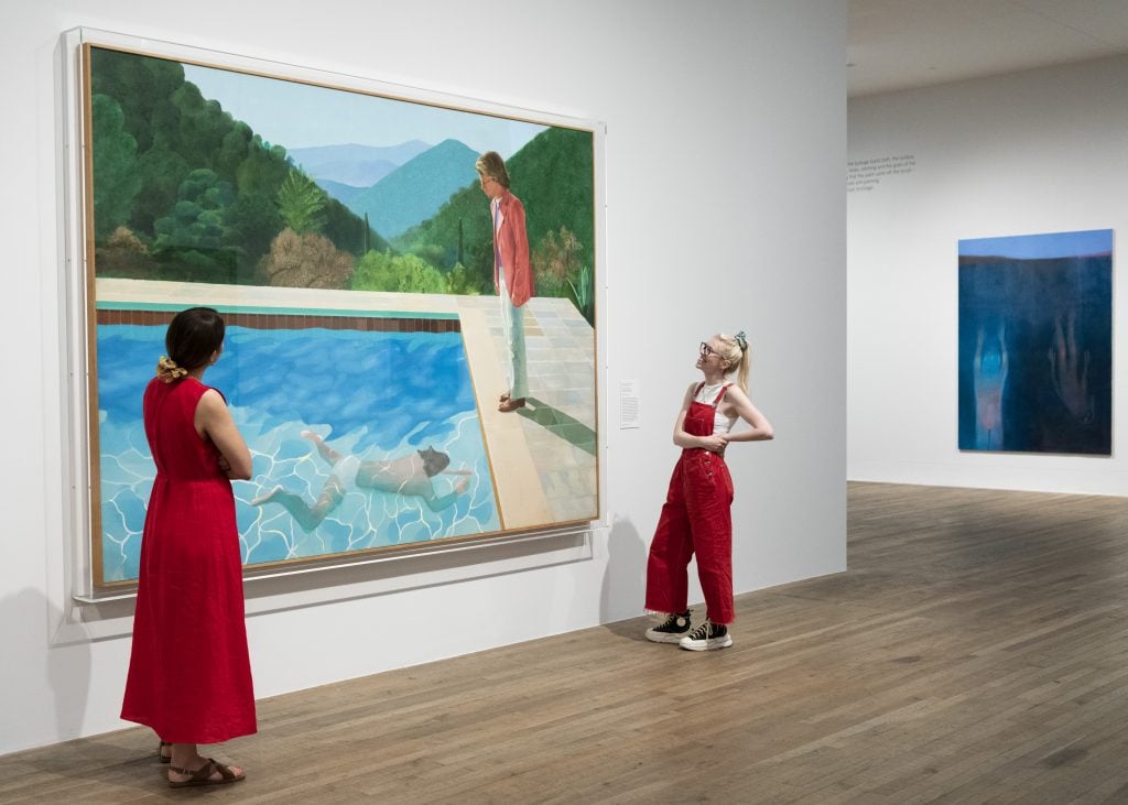 photograph of two women in red outfits viewing a david hockney painting of a man swiming in a pool