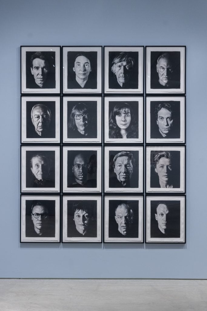 a photograph of 16 framed black and white portraits by David Seidner in a grid of 4x4 on a powder blue wall