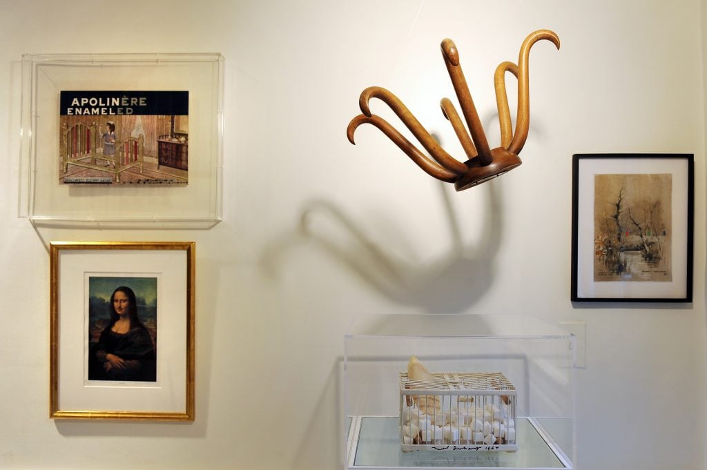 Marcel Duchamp works hanging in a gallery, including a picture of Mona Lisa drawn with a mustache, a hat rack, and a bird cage filled with marble cubes.