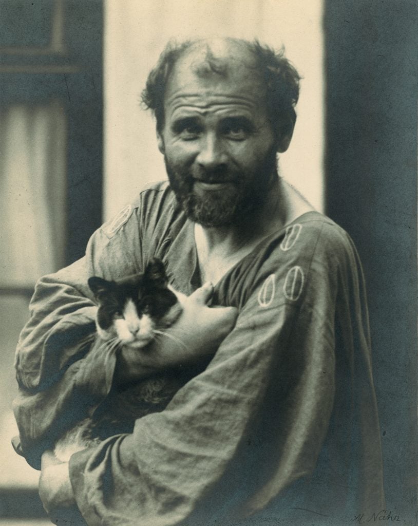Painter Gustav Klint is dressed in a caftan while holding a black and white cat in his arms.