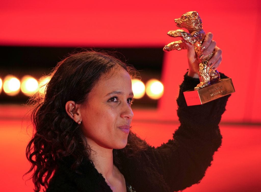 Director Mati Diop stands in front of a red background holding an award that looks like a gold bear
