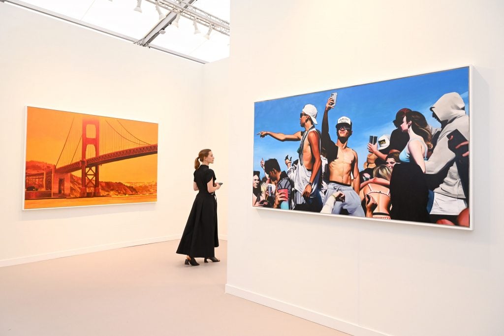 A woman in a black dress passes through an art fair booth between two large photographs, one of the Golden Gate Bridge and another of a group of men.
