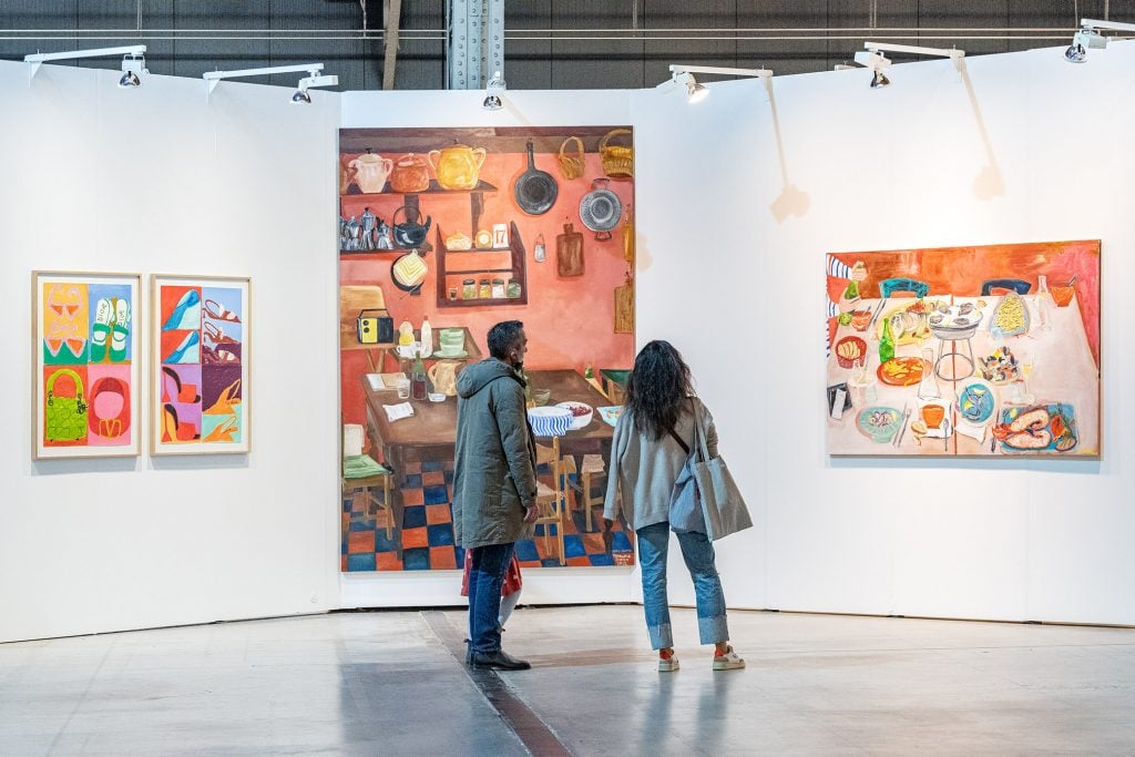 Two people stand in a convention center where three paintings are hung on the walls.
