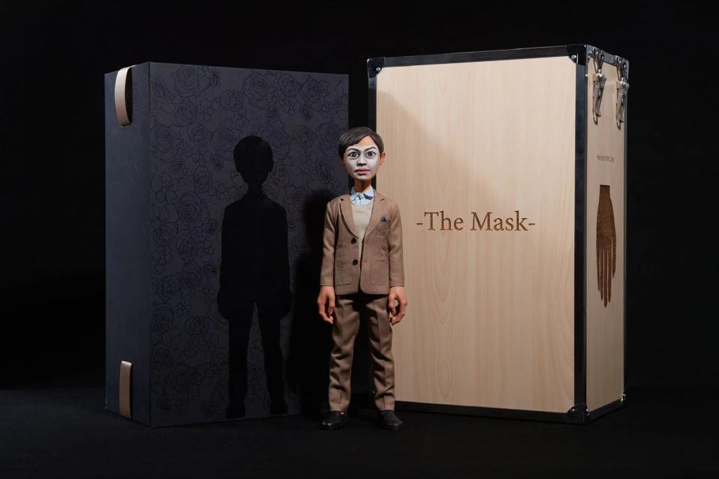 a marionette in a brown suit stands in front of a case labelled the mask. 
