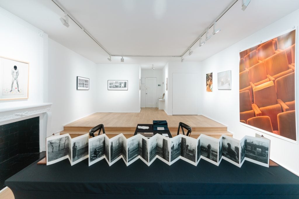 A small white-walled gallery has on its walls a number of photographs in different styles. A black table at front holds an accordion-like book.