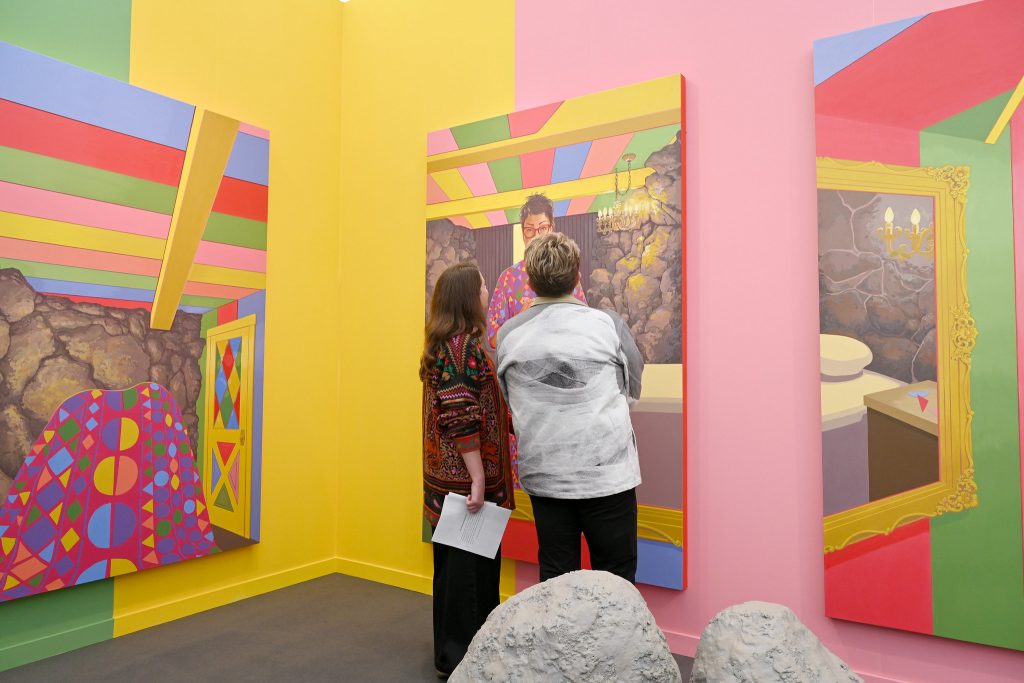 Two people look at colorful paintings in a brightly colored fair booth at Frieze LA.