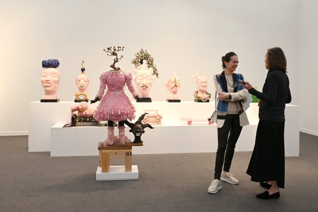 In a color photo, two women stand in front of plinths that display about a dozen pink sculptures that resemble the heads of people. The walls surrounding them are white.