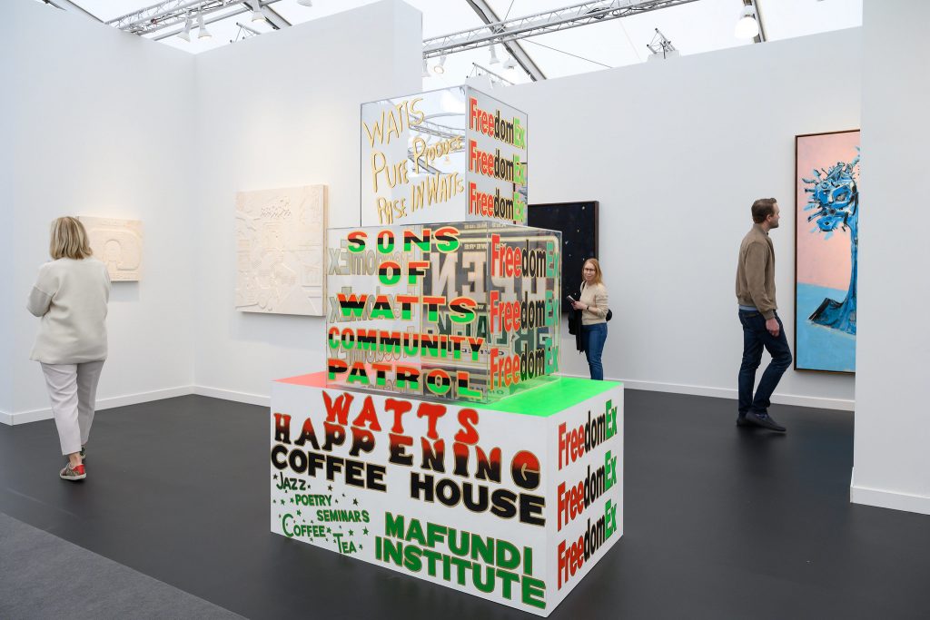 In a color photo, a sculpture of stacked boxes is seen in a white-walled booth with paintings on its walls. The boxy sculpture has phrases on it like "Sons of Watts Community Control" and "Watts Happening Coffee House"