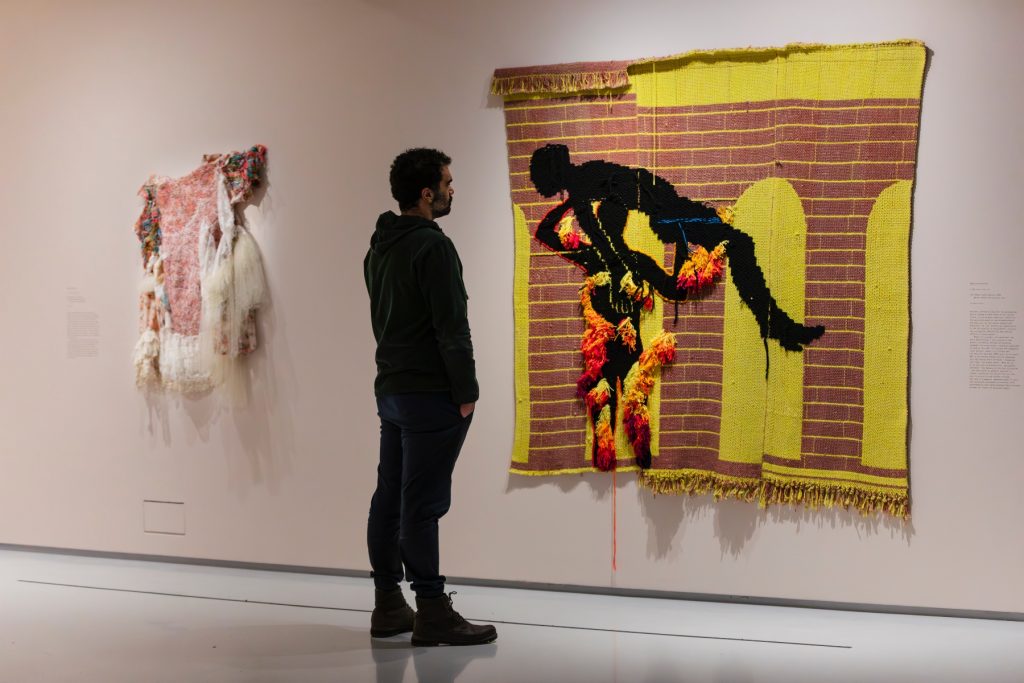 A man looks at a textile on the wall that shows a figure against some arched buildings