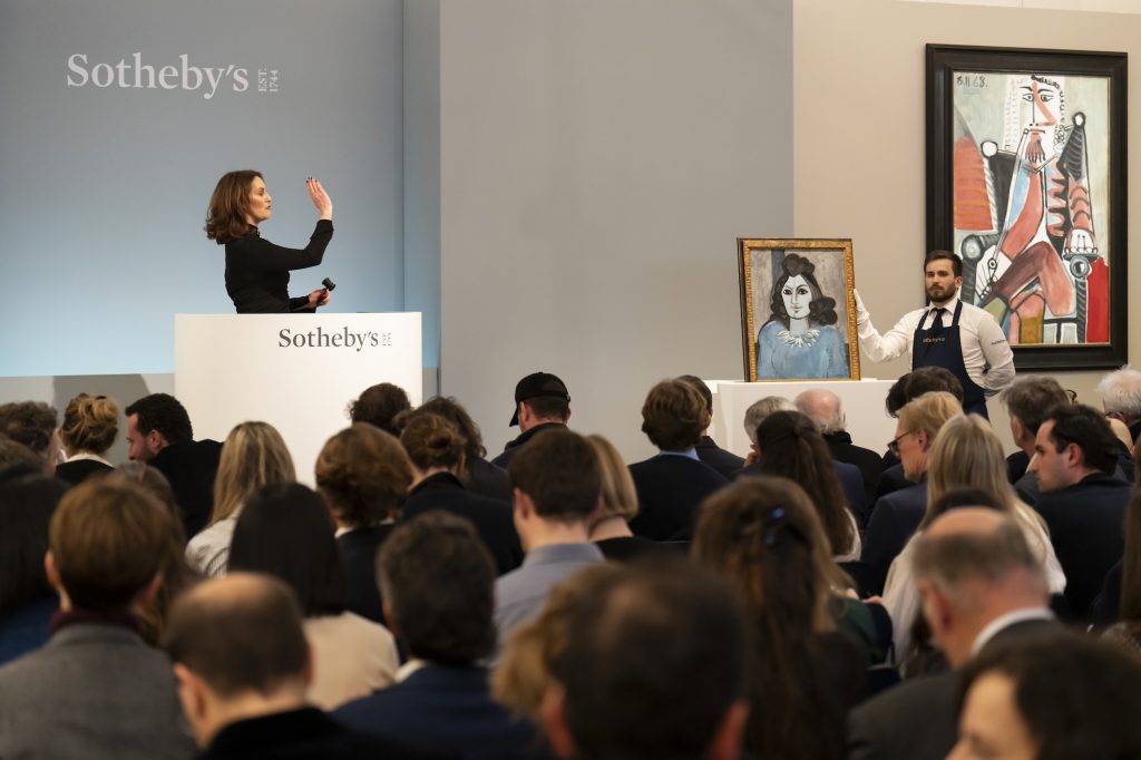 A color photo shows a woman at a podium before a crowd of seated people. Two paintings are to the right of the frame.