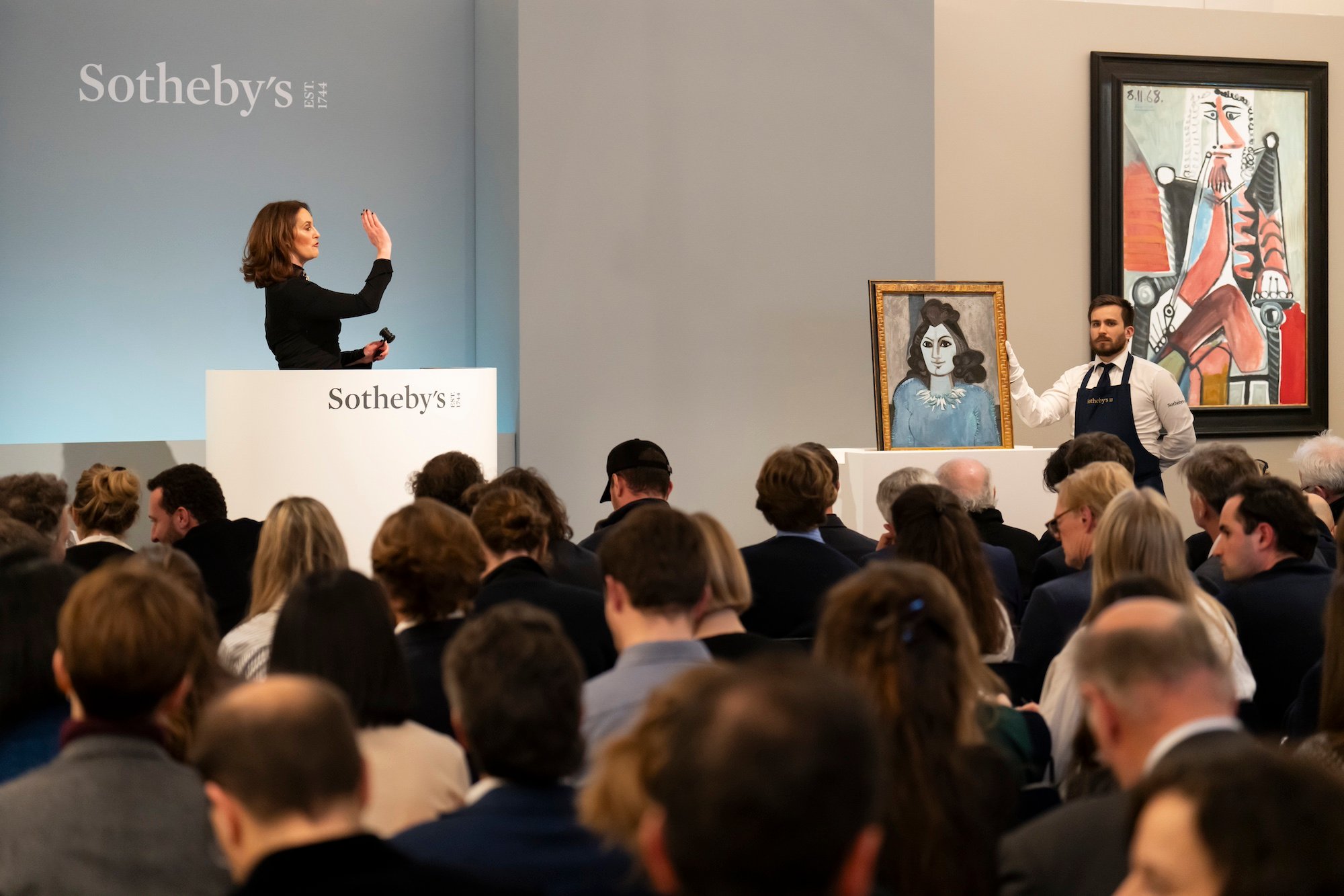 A color photo shows a woman at a podium before a crowd of seated people. Two paintings are to the right of the frame.