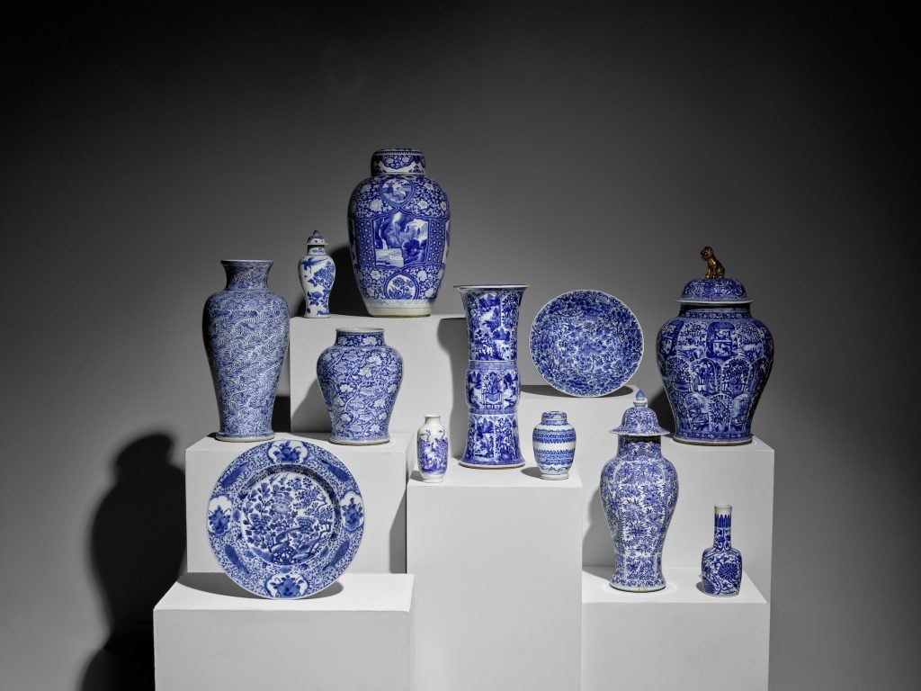 A variety of ornate vases and plates in blue and white sit on white plinths in a dimly lit gallery in a color photograph.